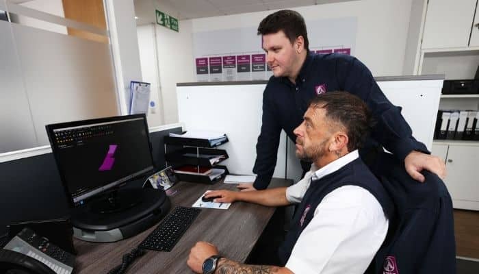 Two men wearing blue and white Avon Group uniform discuss a CAD drawing on a computer in an Avon Group company office